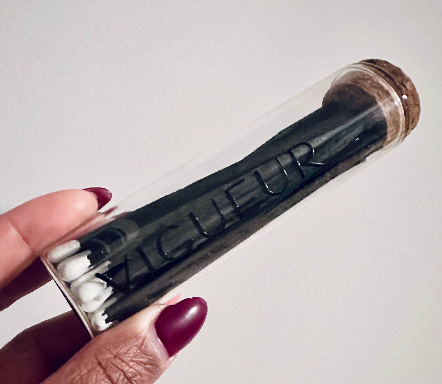Person holding a glass vial filled with matches labeled 'Vigueur' 