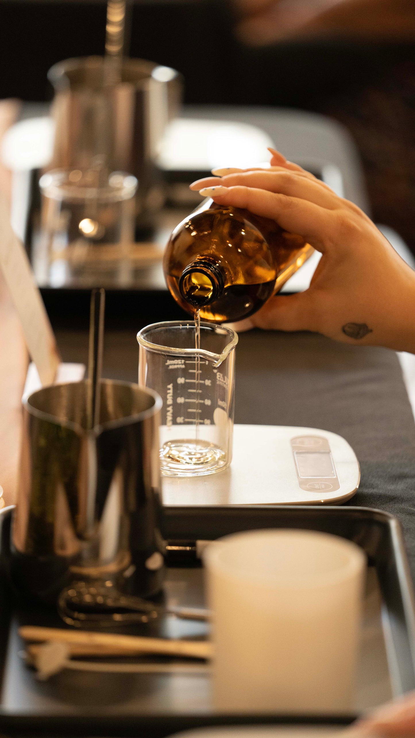 A person's hands pouring wax into a glass candle jar on a table with various other candle making supplies like wicks and tools visible for candle-making workshop.