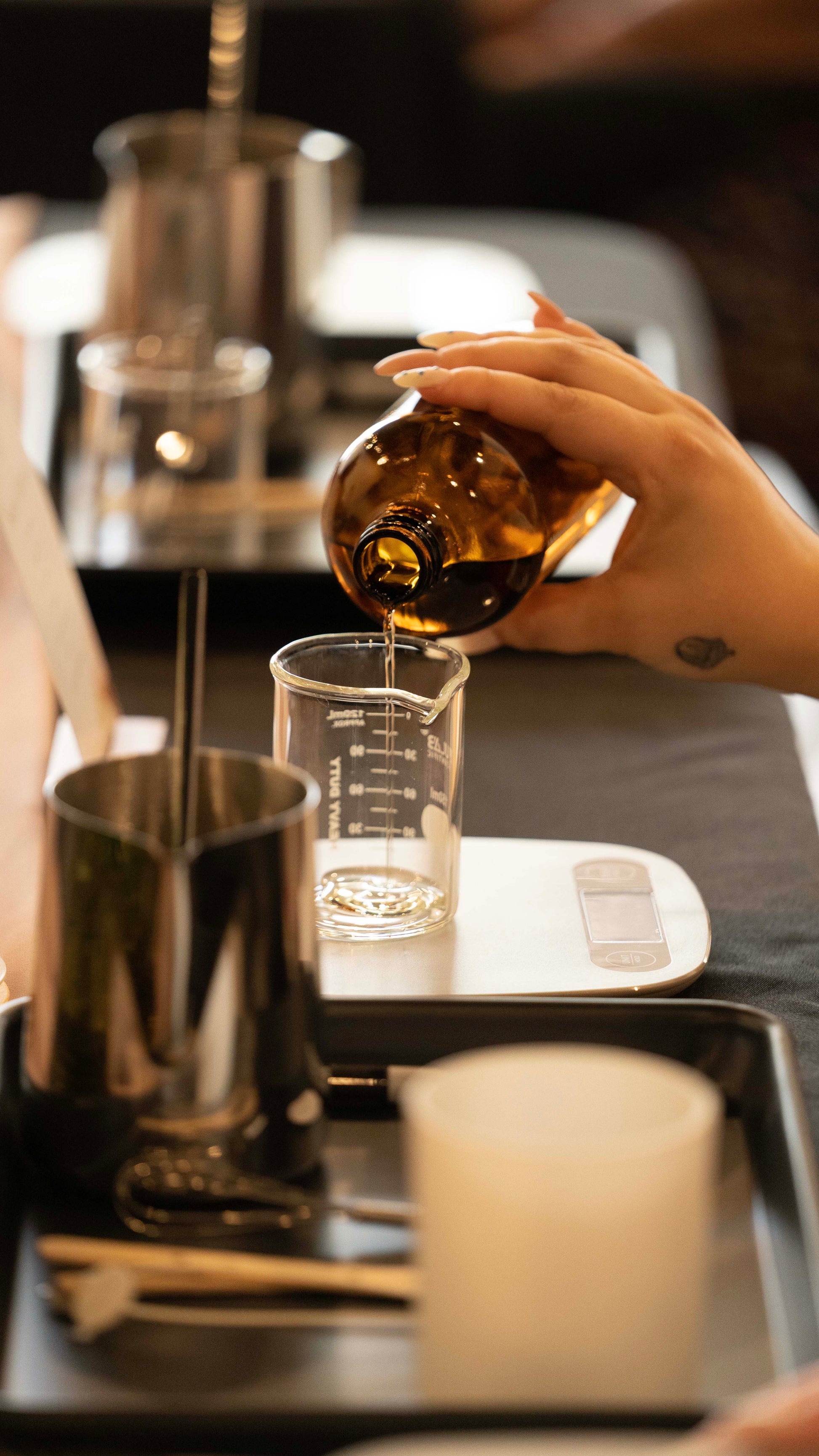 A person's hands pouring wax into a glass candle jar on a table with various other candle making supplies like wicks and tools visible for candle-making workshop.